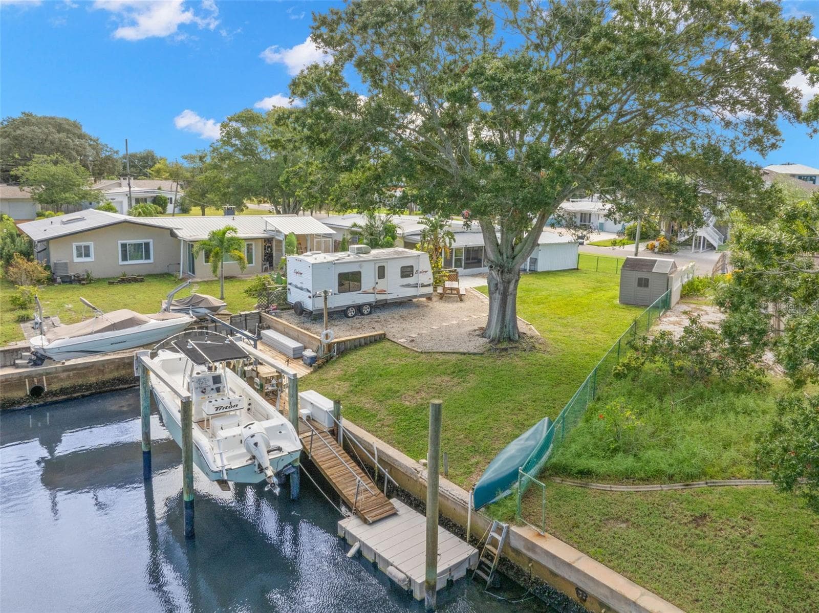 An aerial view showcases the property’s waterfront lifestyle, with a 10,000-pound boat lift, wooden dock and floating dock along the canal, plus generous lawn space, RV parking, and mature shade trees just steps from the water. - 332 WESTWINDS DR, PALM HARBOR
