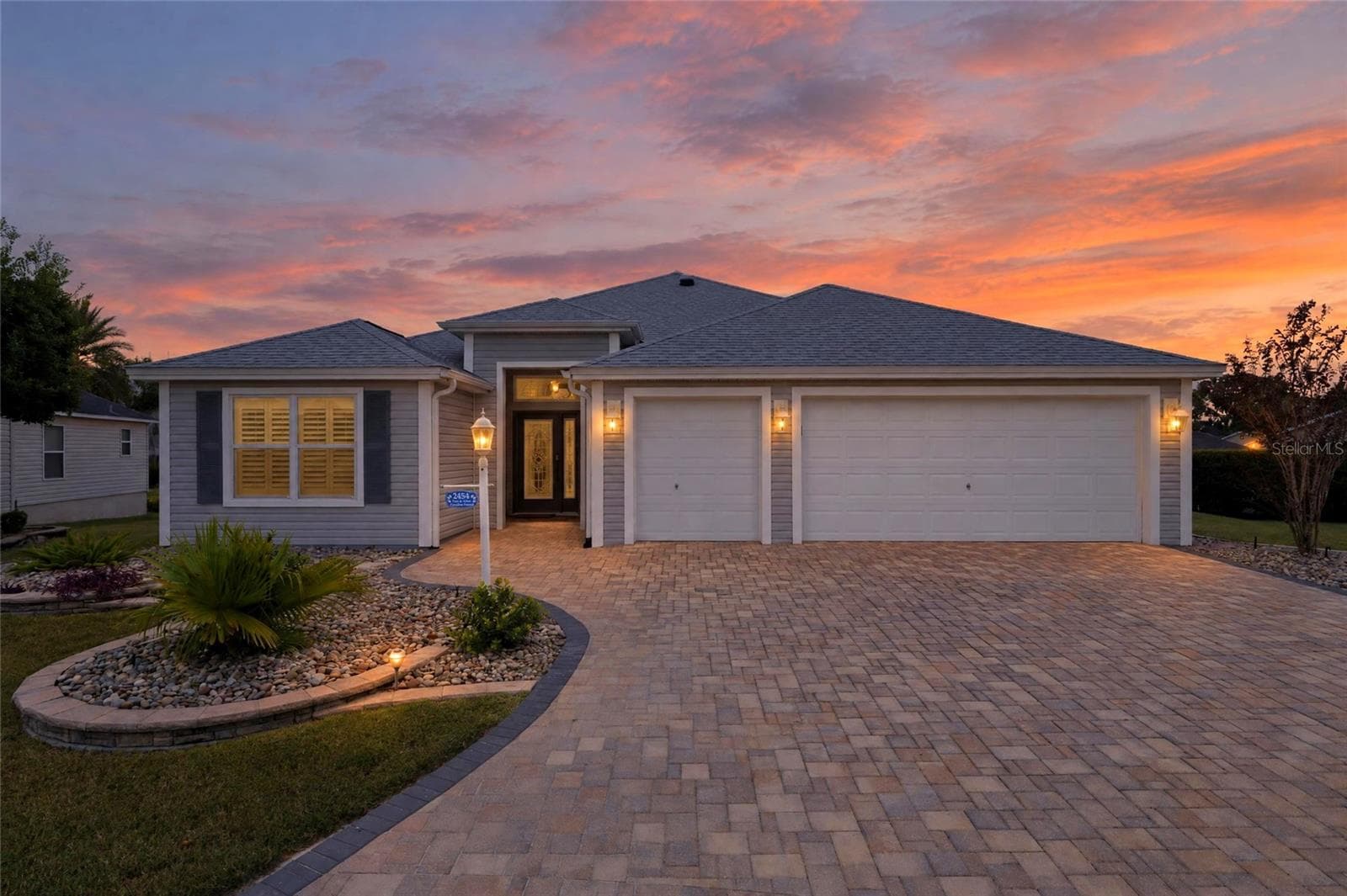 A glowing Florida sunset showcases this home’s beautiful landscaping, sweeping paver driveway, and welcoming curb appeal. - 2454 MORNING DOVE LN, THE VILLAGES