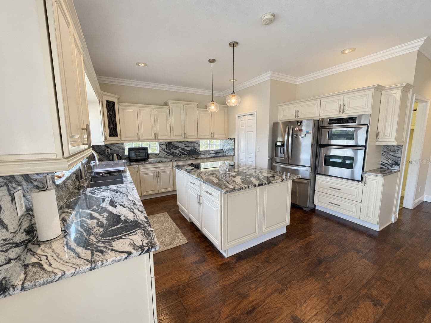 Spacious kitchen featuring cream cabinetry, striking stone countertops and matching backsplash, a central island with an electric cooktop, and stainless steel appliances, including double wall ovens. A large window above the sink overlooks the backyard and water. - 11117 ANCIENT FUTURES DR, TAMPA