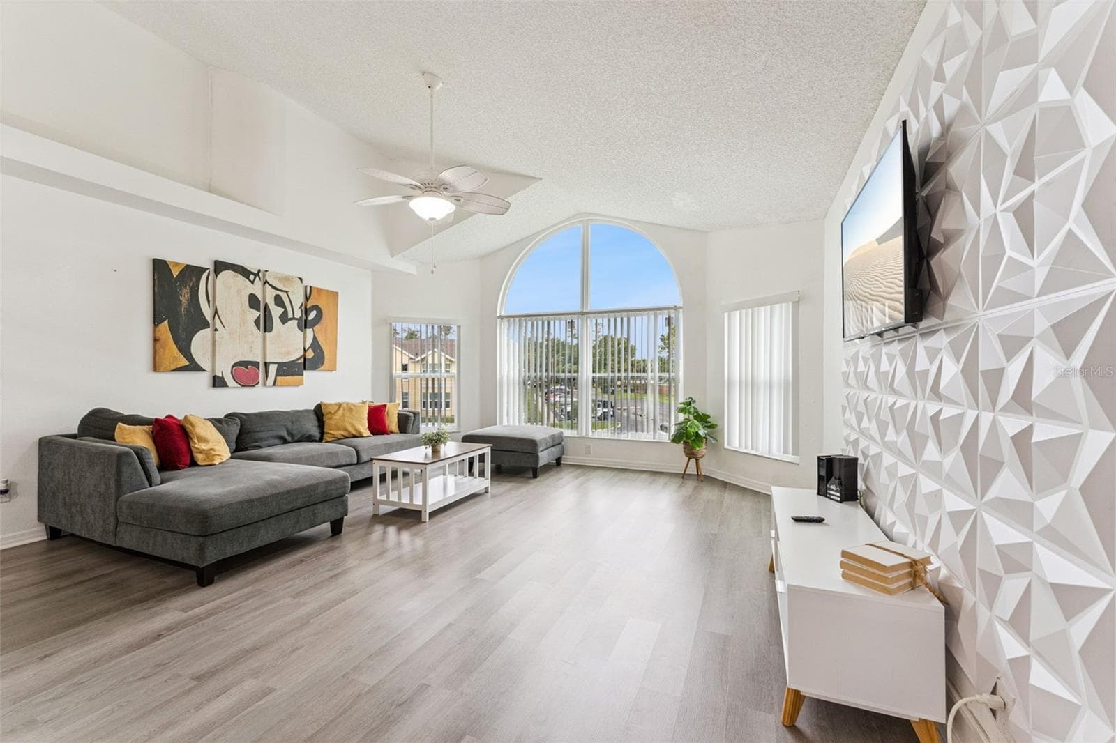 Living Room: Sun-drenched living room boasting vaulted ceilings, a geometric 3D feature wall, wood-look plank flooring, and a large architectural arched window - 5017 LAGUNA BAY CIR #100, KISSIMMEE