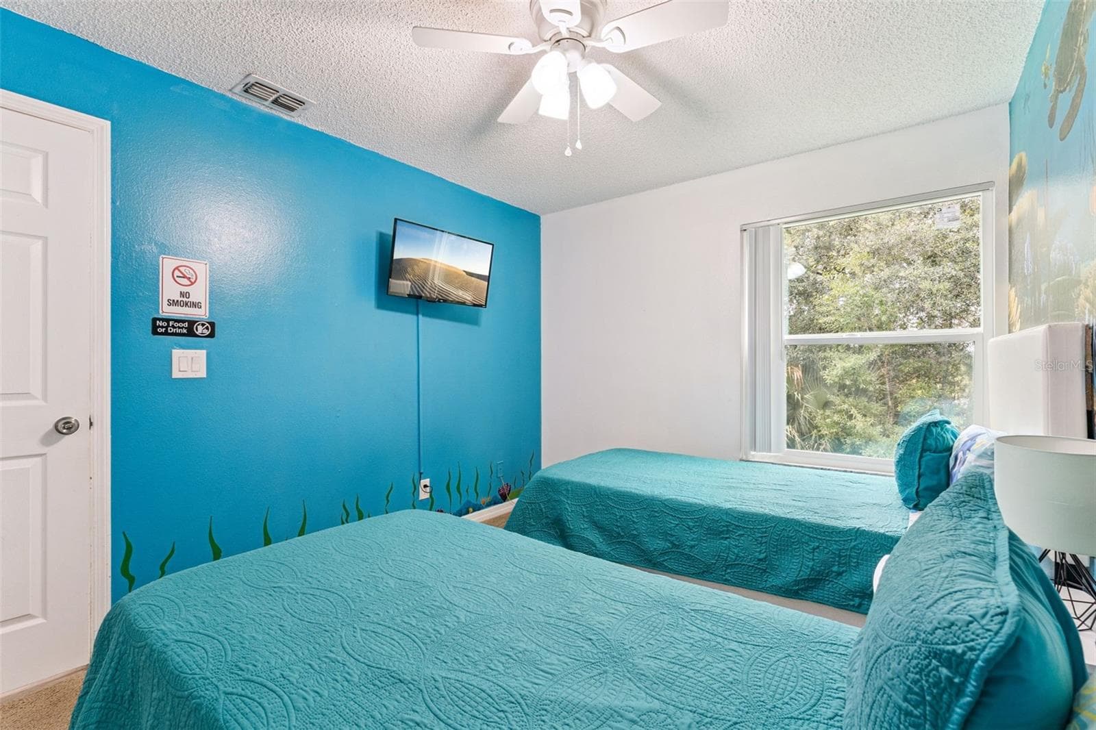 Ocean-Themed Guest Bedroom: Bright guest bedroom featuring vibrant ocean blue accent walls, dual twin beds, a ceiling fan, and wall-mounted TV. - 5017 LAGUNA BAY CIR #100, KISSIMMEE