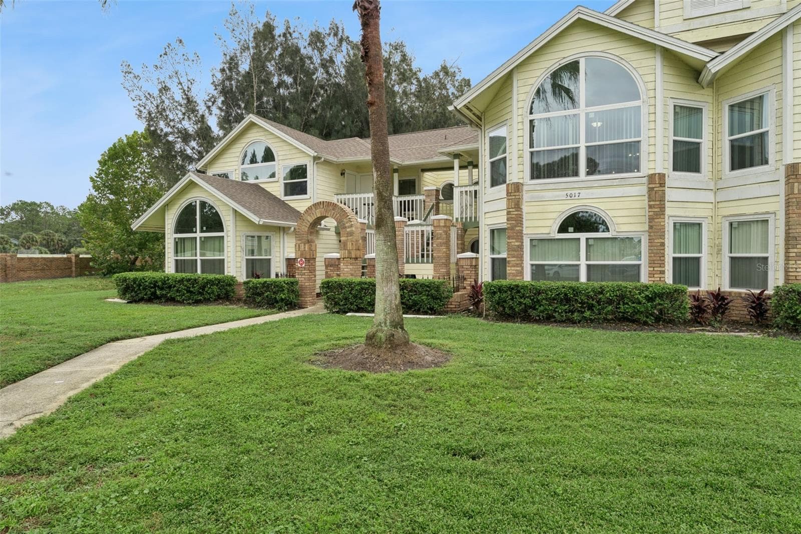 Front Walkway & Entry: Paver walkway leading up to the brick entry archway, framed by mature palm trees and green space. - 5017 LAGUNA BAY CIR #100, KISSIMMEE