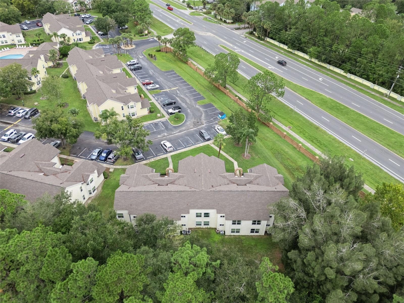 Aerial View of Rear & Grounds: Elevated aerial angle looking over mature tree tops toward the building rear, parking facilities, and nearby main road - 5017 LAGUNA BAY CIR #100, KISSIMMEE
