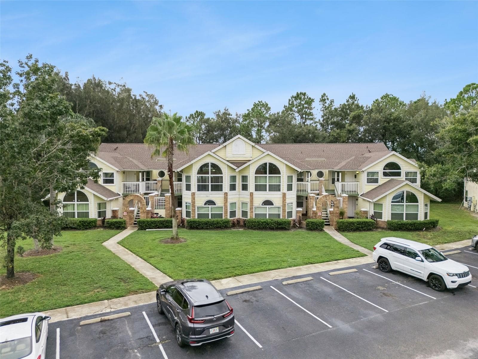 Front Elevation View: Low-angle drone photo showcasing the full building exterior, grand architectural arched windows, entry staircases, and front parking lot. - 5017 LAGUNA BAY CIR #100, KISSIMMEE