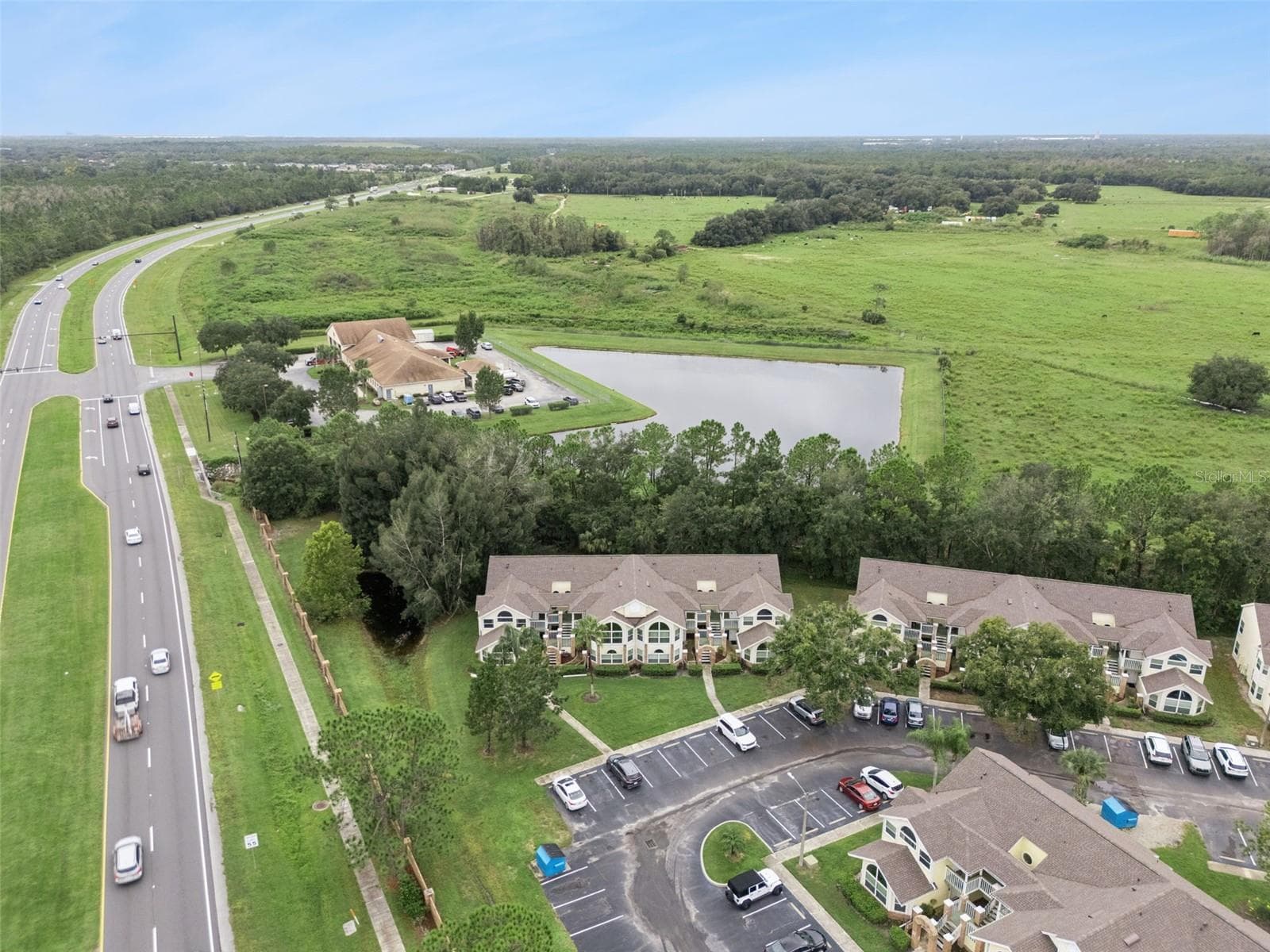 Community & Pond Overview: Broad aerial panorama displaying the condominium grounds, nearby water feature, surrounding open green spaces, and adjacent thoroughfare. - 5017 LAGUNA BAY CIR #100, KISSIMMEE