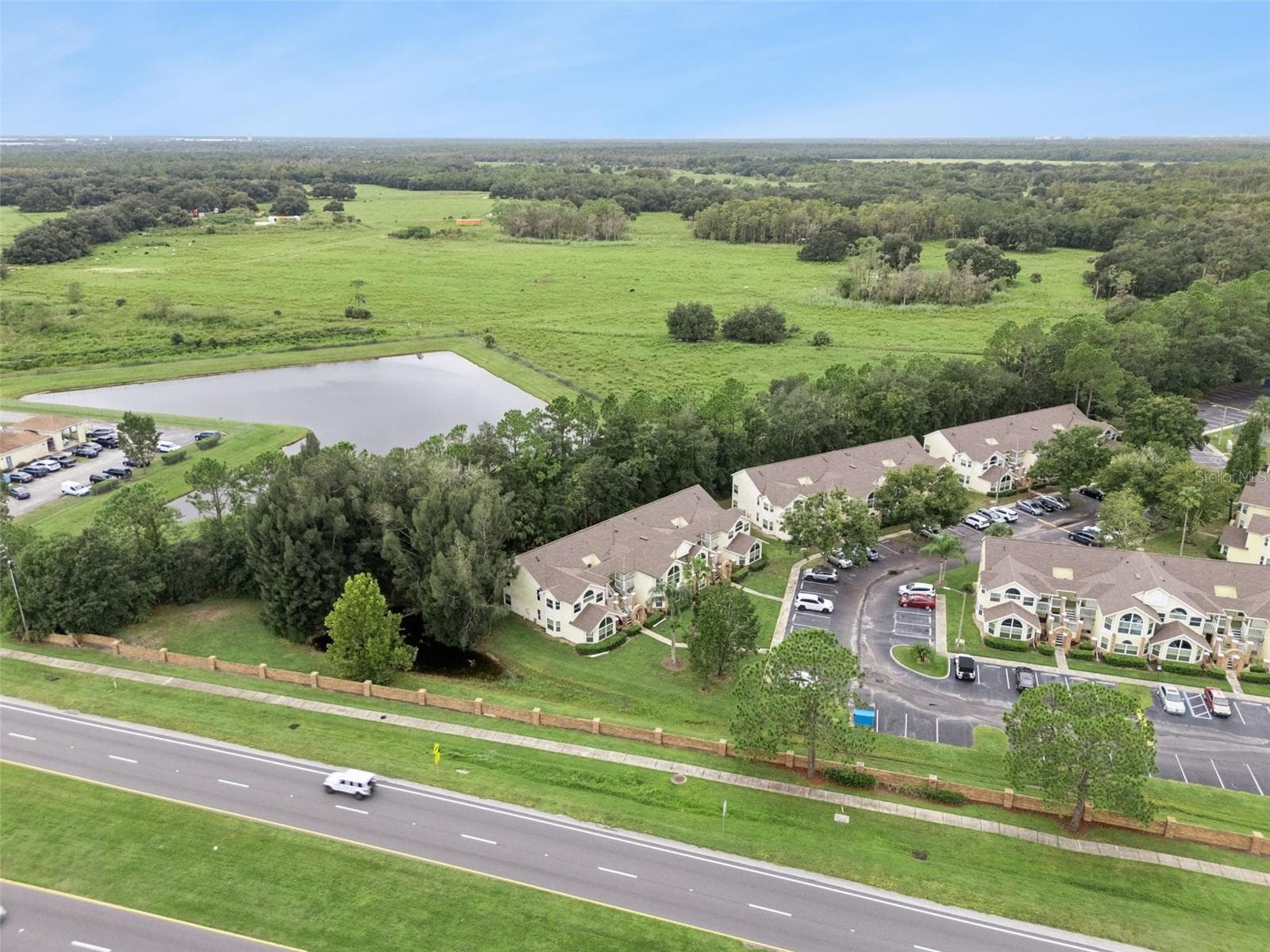 Water Feature & Open Fields Aerial: Wide aerial perspective featuring the community's adjacent tranquil pond, wooded buffer line, and rolling green backdrop. - 5017 LAGUNA BAY CIR #100, KISSIMMEE