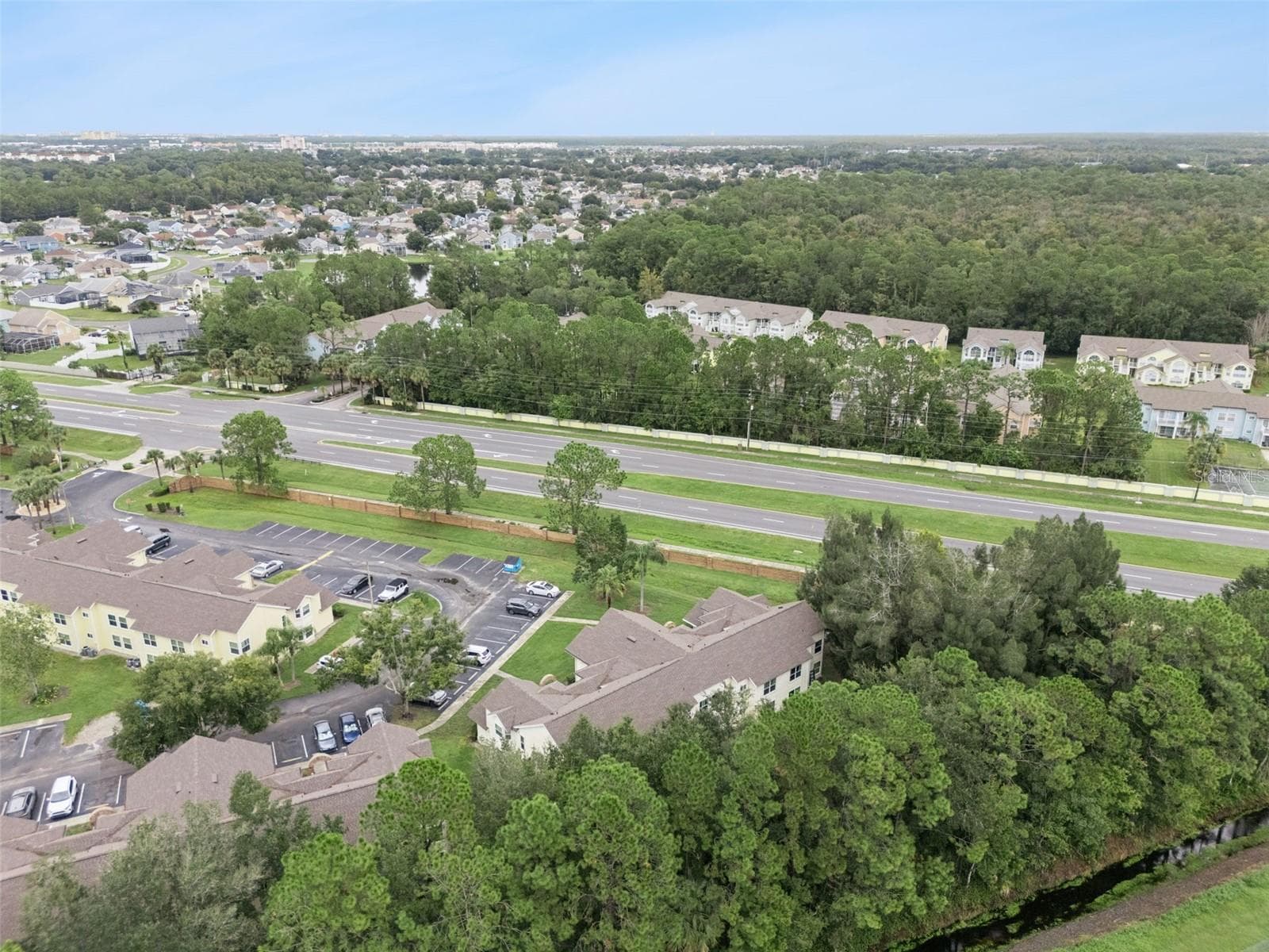 High-Angle Regional Aerial: Elevated drone view displaying the building nestled beside a lush pine tree buffer, showing parking access, surrounding neighborhoods, and convenient proximity to major roadways. - 5017 LAGUNA BAY CIR #100, KISSIMMEE
