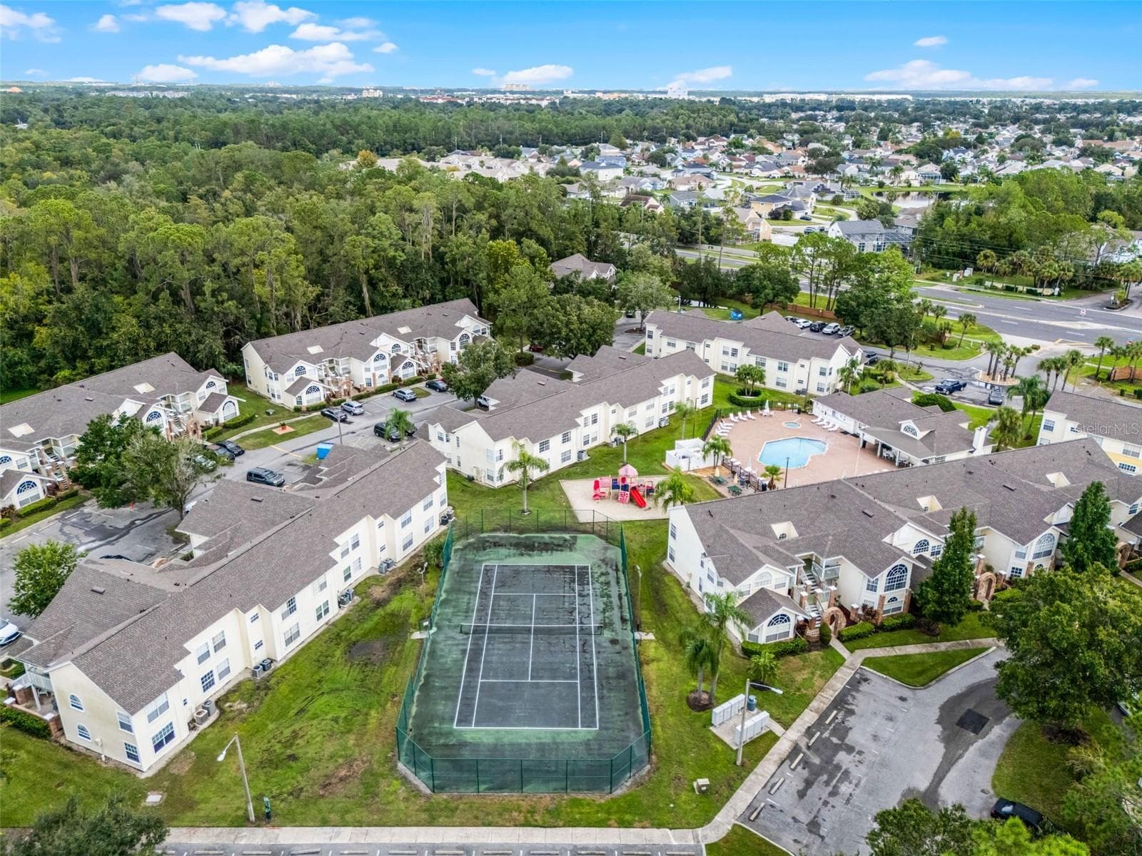 Community Amenities Aerial: High-angle drone shot displaying key community features, including the fenced tennis court, children's playground, resort pool, clubhouse, and surrounding residential buildings. - 5017 LAGUNA BAY CIR #100, KISSIMMEE