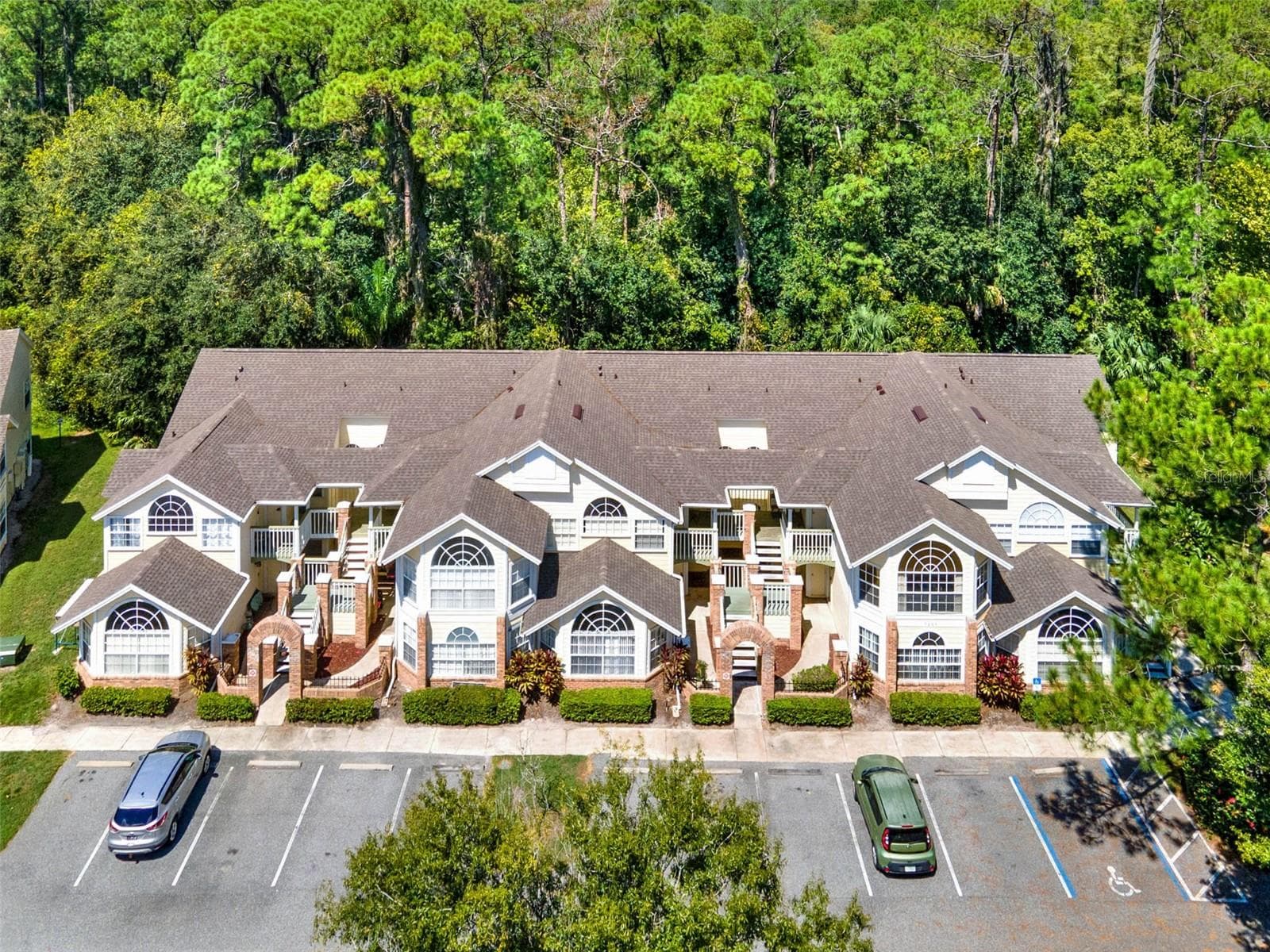 uilding Exterior Aerial: Direct front aerial perspective showcasing the unit building, parking spaces, brick entry archways, and dense wooded tree canopy behind. - 5017 LAGUNA BAY CIR #100, KISSIMMEE