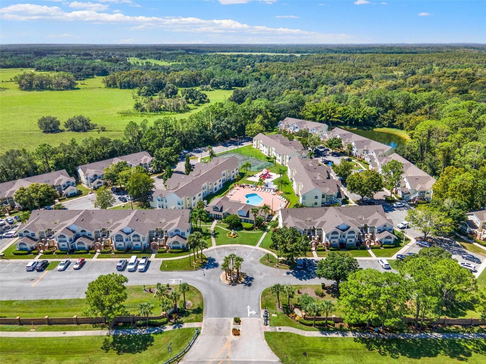 Community Entrance & Layout Aerial: Broad aerial view capturing the main entrance roundabout, central pool deck, clubhouse, tennis court, and residential layout. - 5017 LAGUNA BAY CIR #100, KISSIMMEE