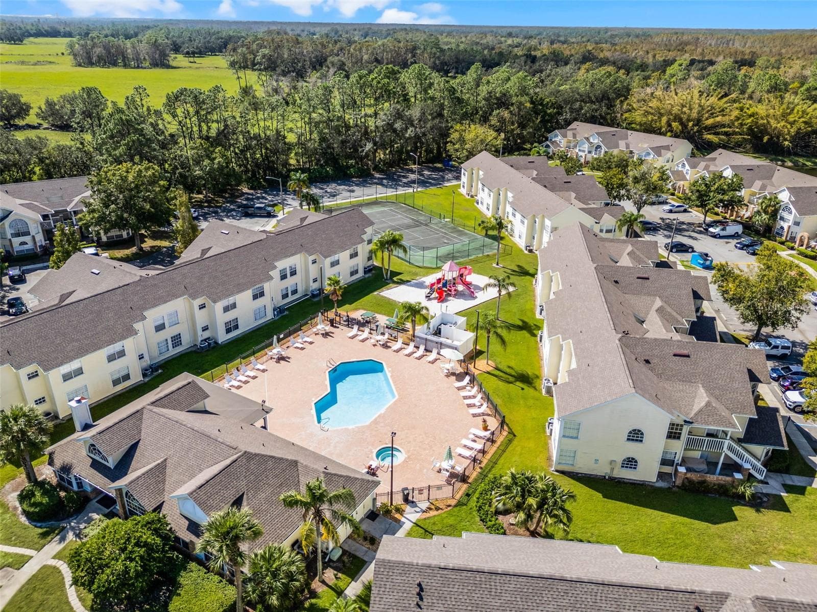 Amenity Hub Aerial: Elevated aerial view highlighting the central pool deck, clubhouse, sand playground, tennis court, and surrounding residential buildings set against open green spaces. - 5017 LAGUNA BAY CIR #100, KISSIMMEE