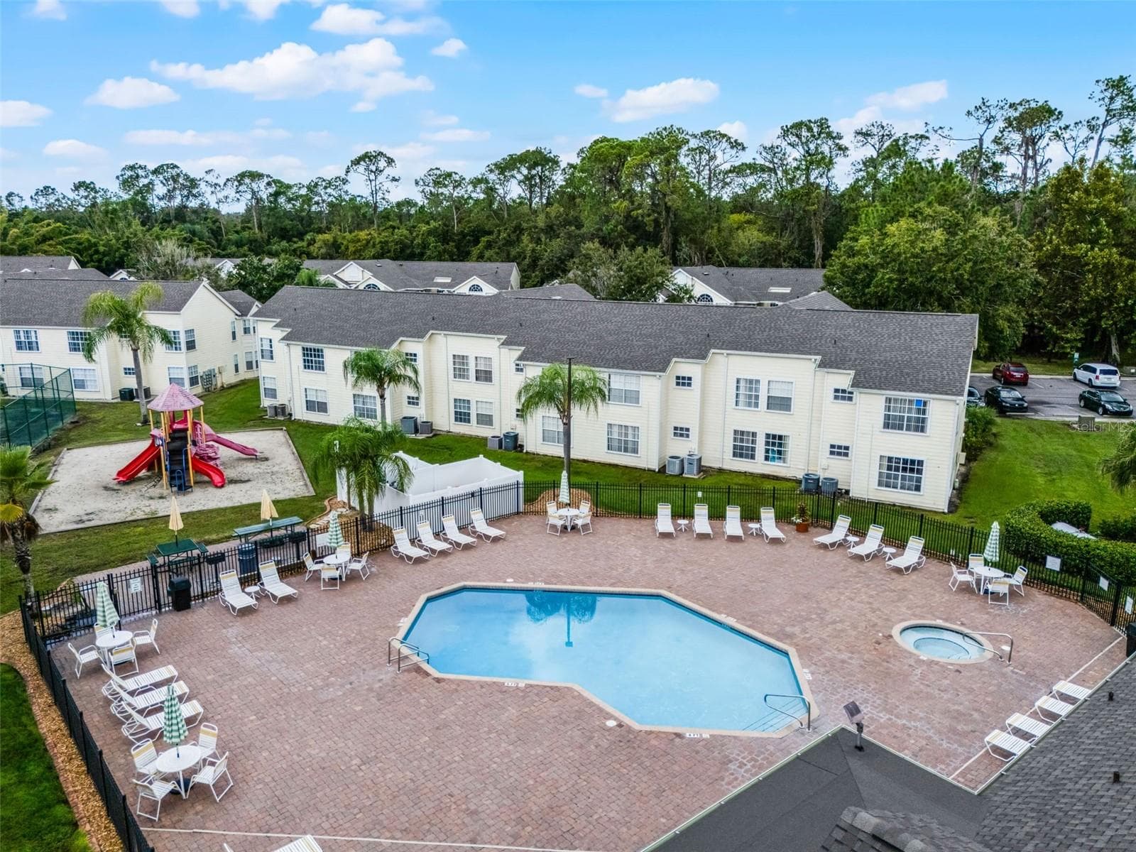 Resort Pool & Playground View: Angled drone view of the paver pool deck, integrated hot tub, lounge seating, fenced perimeter, and adjacent children's playground. - 5017 LAGUNA BAY CIR #100, KISSIMMEE