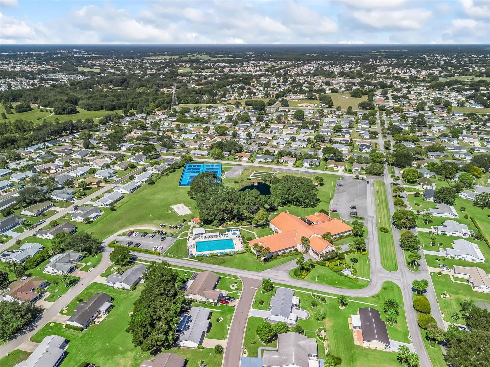This panoramic aerial view showcases Spruce Creek South’s clubhouse and recreation area, including the swimming pool, tennis and pickleball courts, shuffleboard courts, and tree-lined pond. Landscaped grounds and surrounding homes highlight the community’s inviting setting. - 17909 SE 99TH AVE, SUMMERFIELD