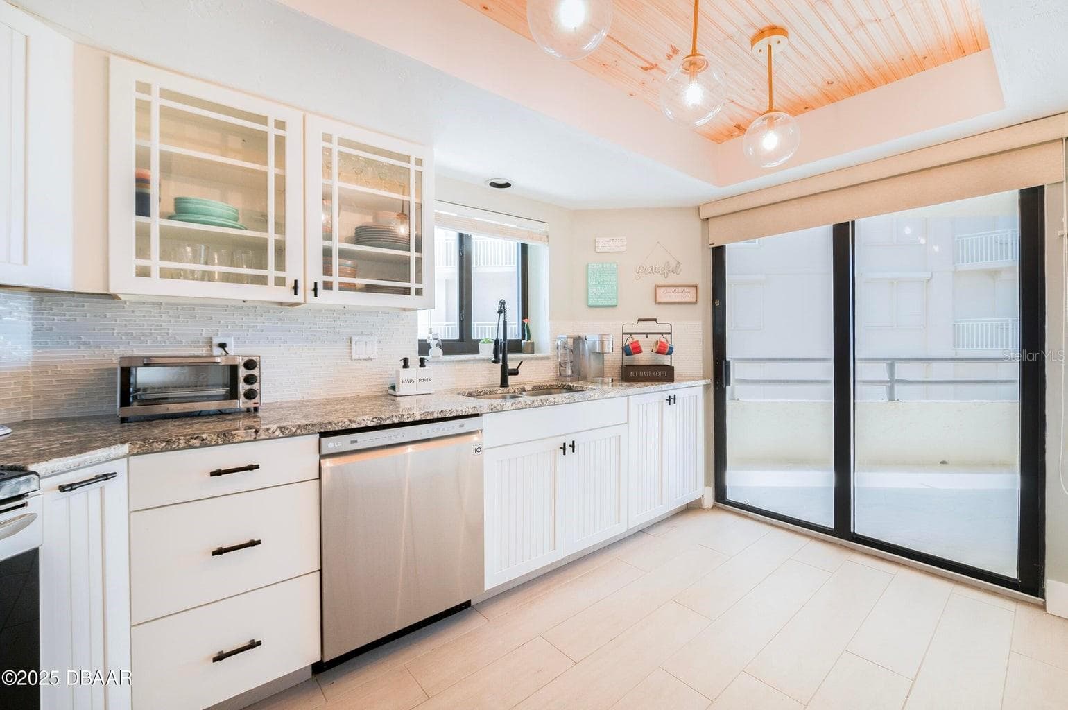 Beautifully appointed kitchen featuring white cabinetry, granite countertops, a stylish tile backsplash, and glass-front upper cabinets. A wood-accented tray ceiling with globe pendant lighting adds warmth and architectural character. - 3855 S ATLANTIC AVE #401, DAYTONA BEACH