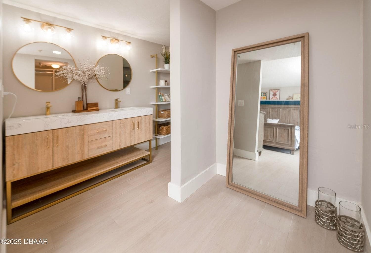 This beautifully updated primary bathroom features a double-sink vanity with wood-tone cabinetry, gold-tone fixtures, round mirrors, and contemporary lighting for a sophisticated coastal look. - 3855 S ATLANTIC AVE #401, DAYTONA BEACH