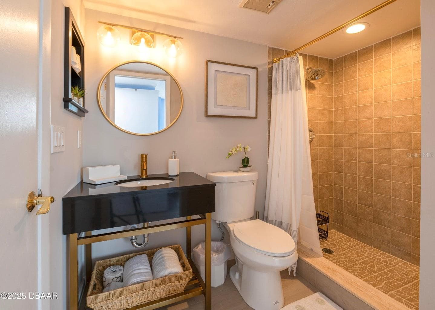 This stylish guest bathroom features a modern black vanity with gold-tone accents, a round mirror, updated lighting, and a tiled walk-in shower with a rainfall showerhead. - 3855 S ATLANTIC AVE #401, DAYTONA BEACH