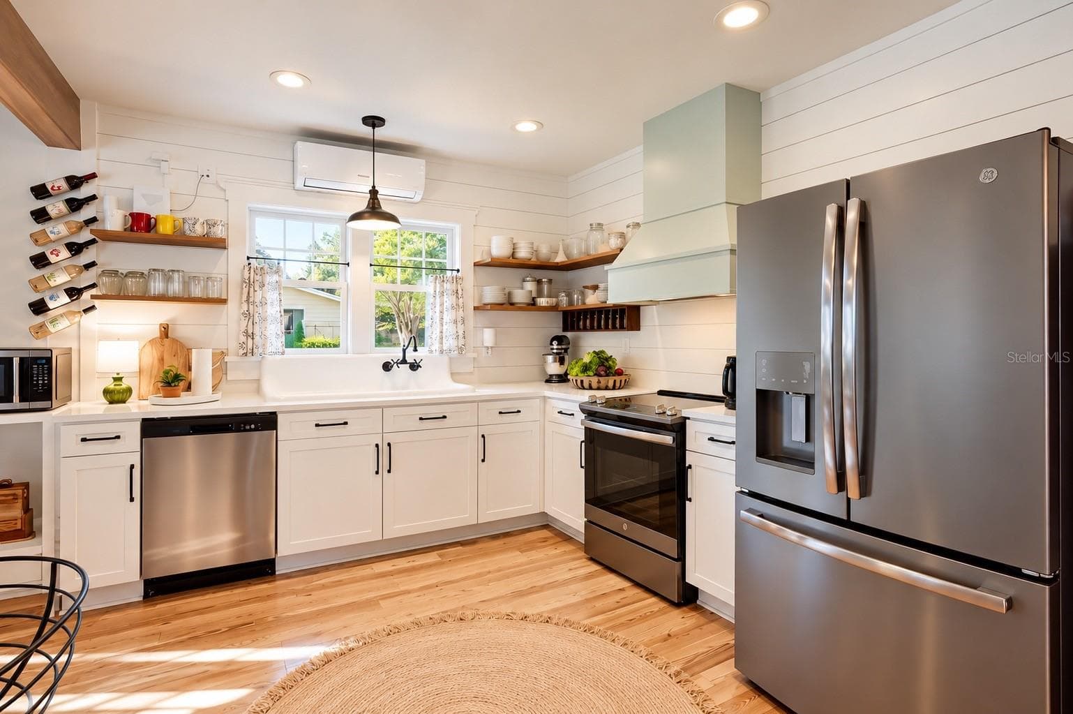 FARMHOUSE-STYLE KITCHEN featuring white shaker cabinetry, shiplap walls, floating wood shelves, gorgeous wood flooring, stainless steel appliances, and an OVERSIZED FARMHOUSE APRON-FRONT SINK with wall-mounted faucet beneath sun-filled windows overlooking the lush backyard, filled with an abundance of EDIBLE TREES, PLANTS, AND HERBS, offering a wonderful opportunity to embrace FARM-TO-TABLE LIVING RIGHT IN YOUR OWN BACKYARD! - 1901 10TH ST, ST CLOUD