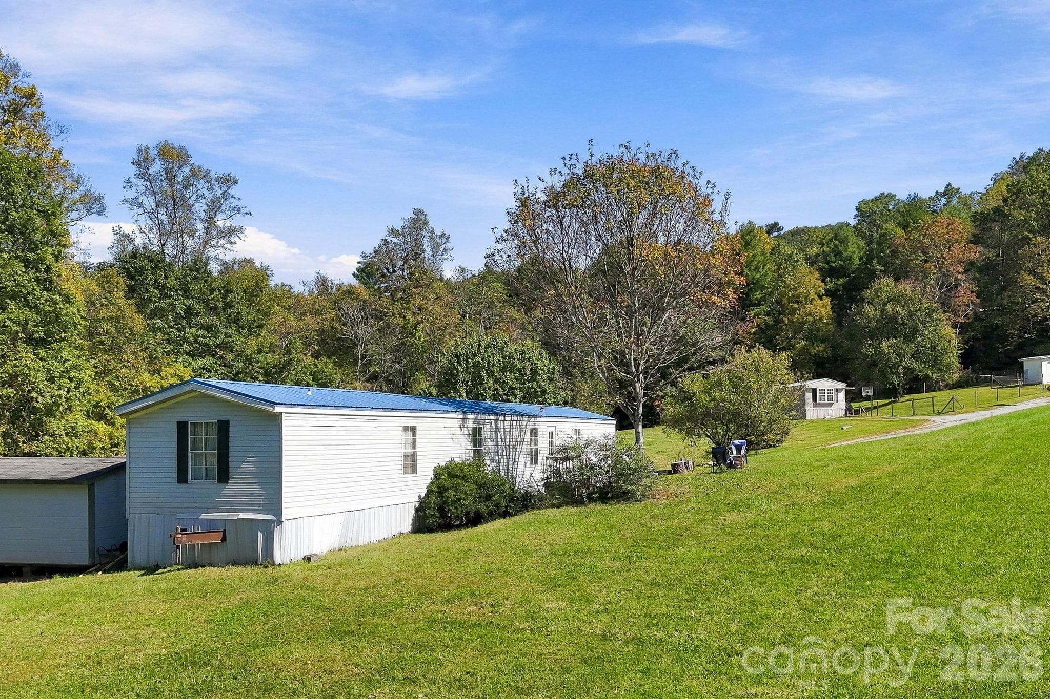 Mobile home with a blue metal roof overlooking a broad grassy lawn, surrounded by mature trees and natural landscaping. - 631 Union Hill Road, Hendersonville