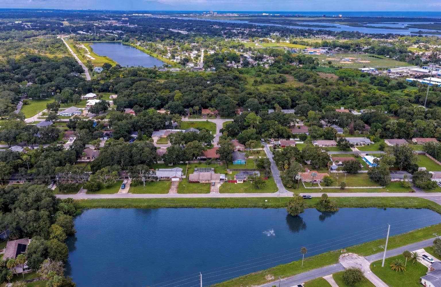High-angle aerial view of the single-story home situated directly across from a peaceful neighborhood lake with a central water fountain. The spacious property features a grassy yard surrounded by mature palm trees and lush local vegetation. - 1101 16TH ST, EDGEWATER