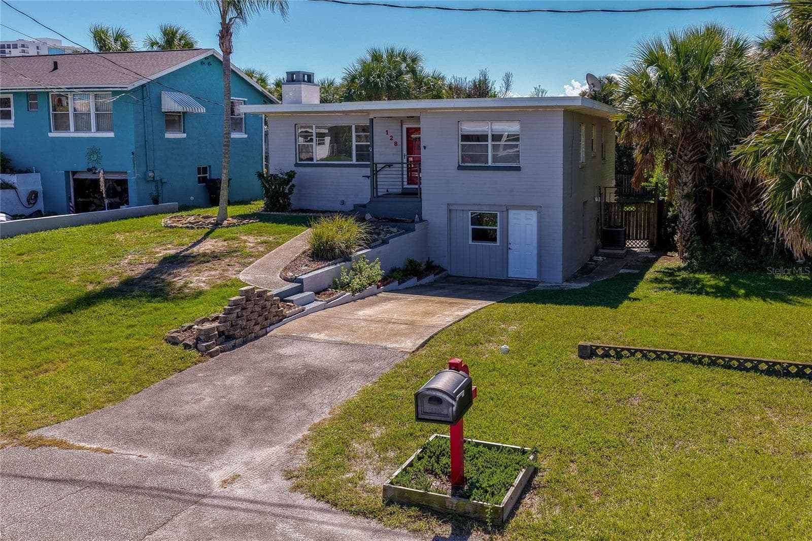 Front exterior perspective of the two-story residence showcasing a paved driveway, a red mailbox fixture, and mature palm trees. The sloped front yard is framed by stone retaining wall accents. - 128 MINERVA RD, DAYTONA BEACH