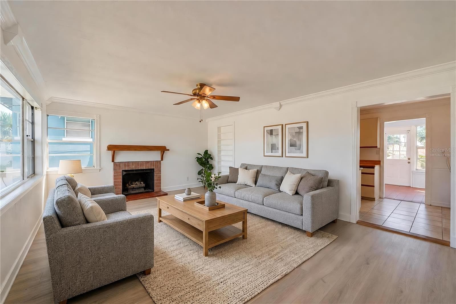Virtually Staged - Spacious living area showcasing light LVP flooring, crown molding, and a charming brick fireplace with a wooden mantel. Large windows allow plenty of natural light, and an open doorway leads toward the kitchen and sunroom areas. - 128 MINERVA RD, DAYTONA BEACH