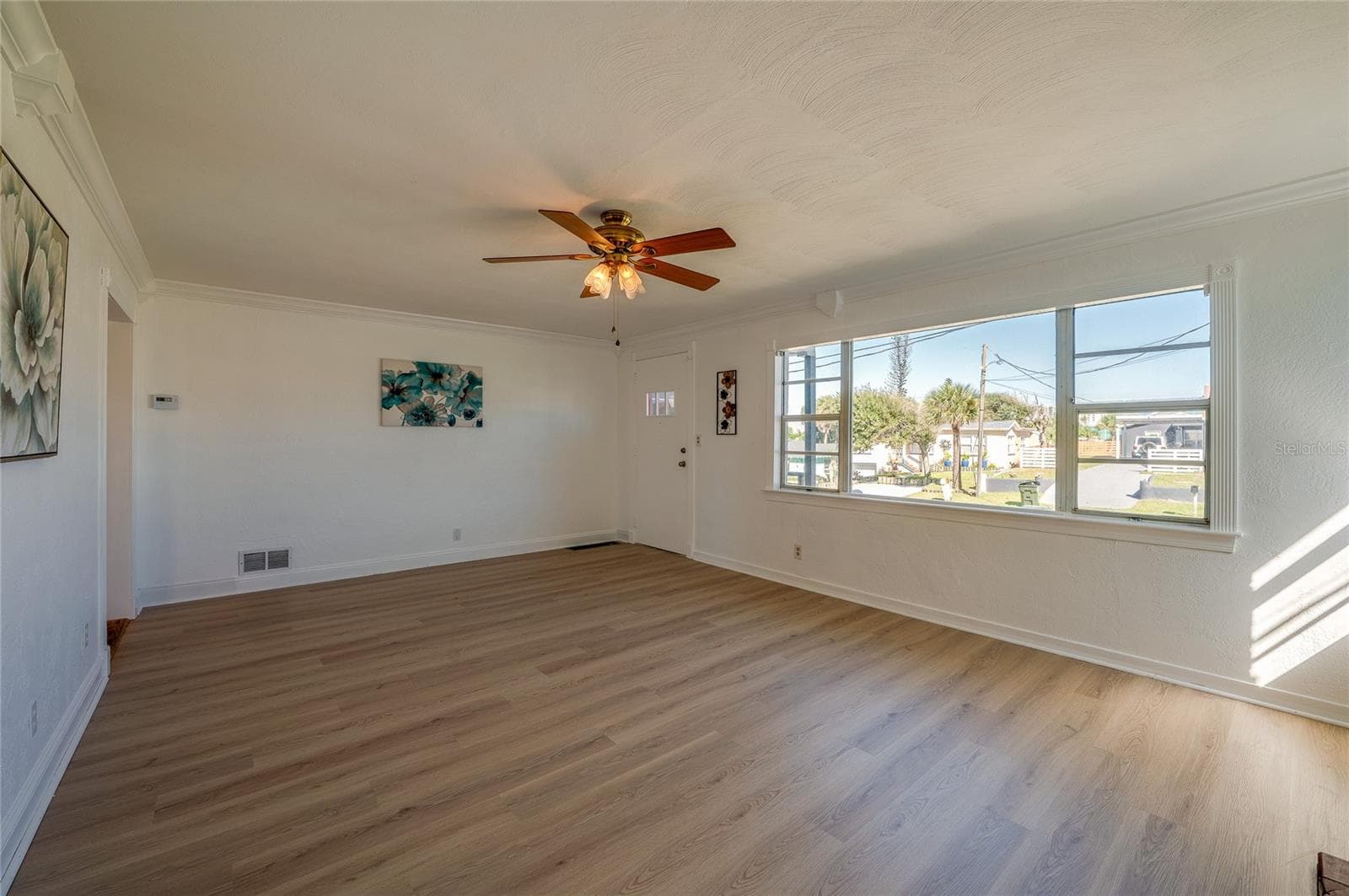 Bright living room highlighting smooth neutral walls, a ceiling fan with light fixtures, and a expansive front window overlooking the neighborhood. A side doorway connects seamlessly to the rest of the home's interior layout. - 128 MINERVA RD, DAYTONA BEACH