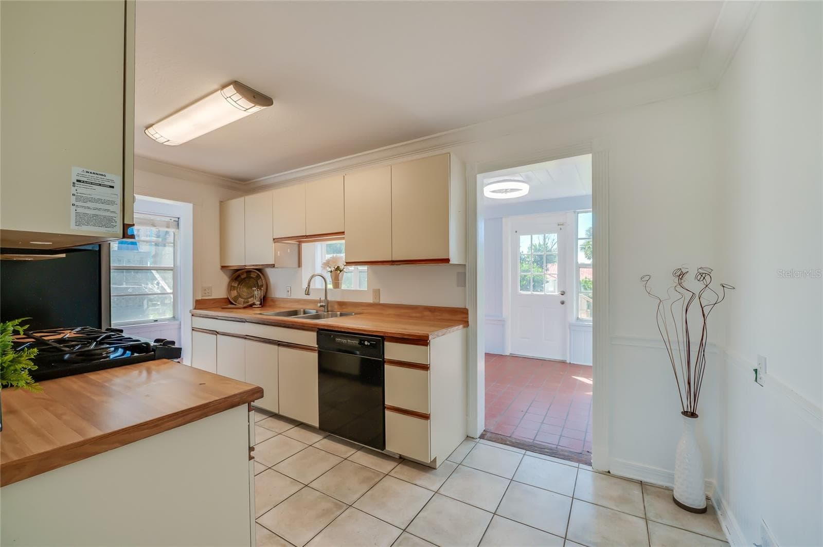 Functional kitchen featuring butcher block-style countertops, classic cabinetry, tile flooring, and integrated black appliances. An adjacent open doorway provides direct access to a sunlit room with terracotta tile floors. - 128 MINERVA RD, DAYTONA BEACH