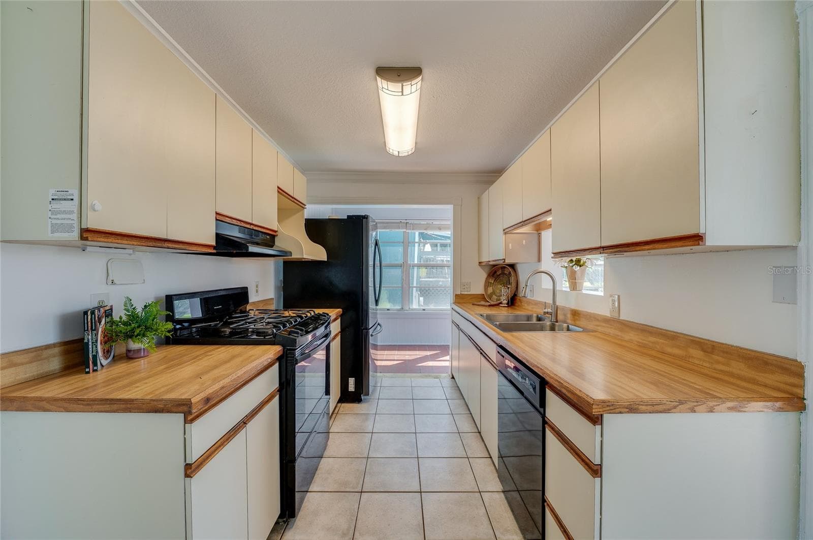 Galley-style kitchen design equipped with dual-sided cabinetry, warm wood-toned countertops, a double-basin sink, and overhead light fixtures. The walkthrough layout opens up directly to the sunroom beyond. - 128 MINERVA RD, DAYTONA BEACH