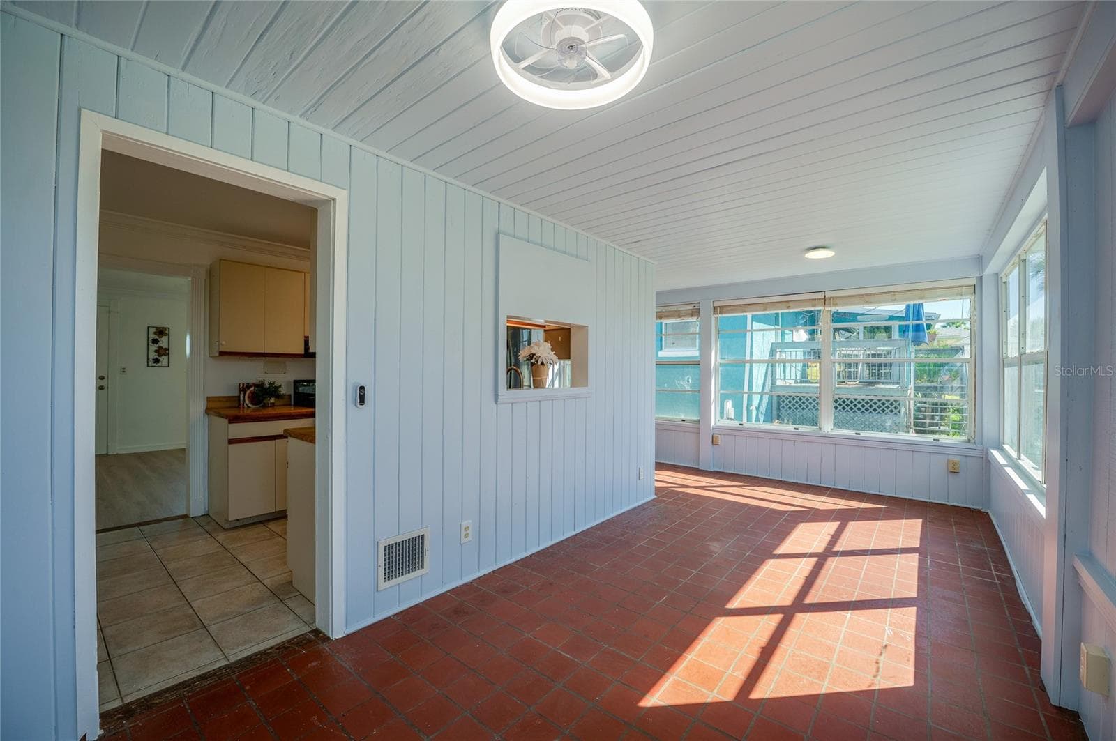 Dinning area featuring light blue wood-paneled walls, warm terracotta floor tiles, and abundant wrap-around windows. A passthrough window connects back to the kitchen for added convenience. - 128 MINERVA RD, DAYTONA BEACH