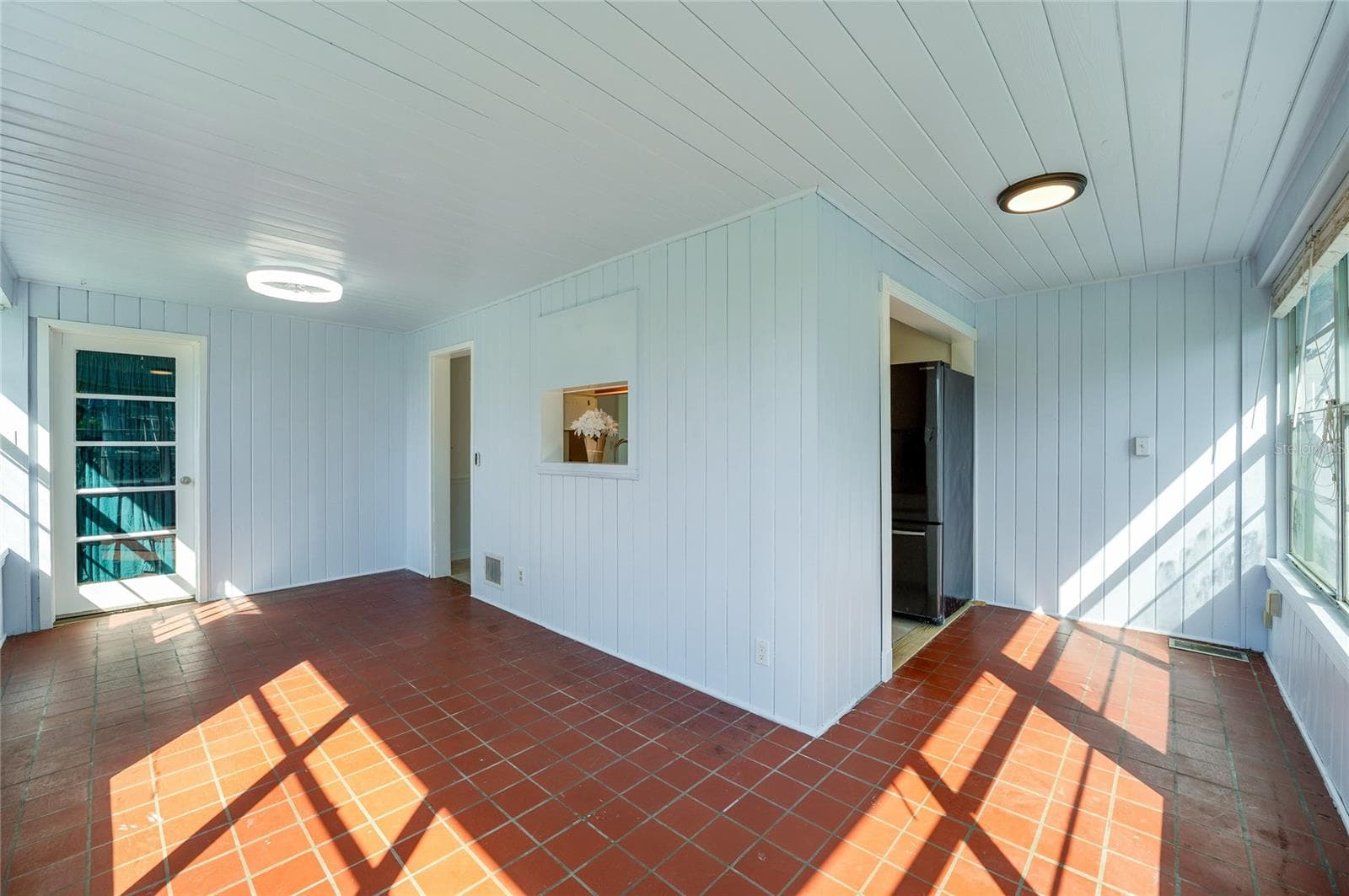 Sun-filled dining area and breakfast nook showcasing red tile flooring, modern flush-mount lighting, and white paneled walls. Large windows fill the space with natural light while direct door access leads outdoors. - 128 MINERVA RD, DAYTONA BEACH