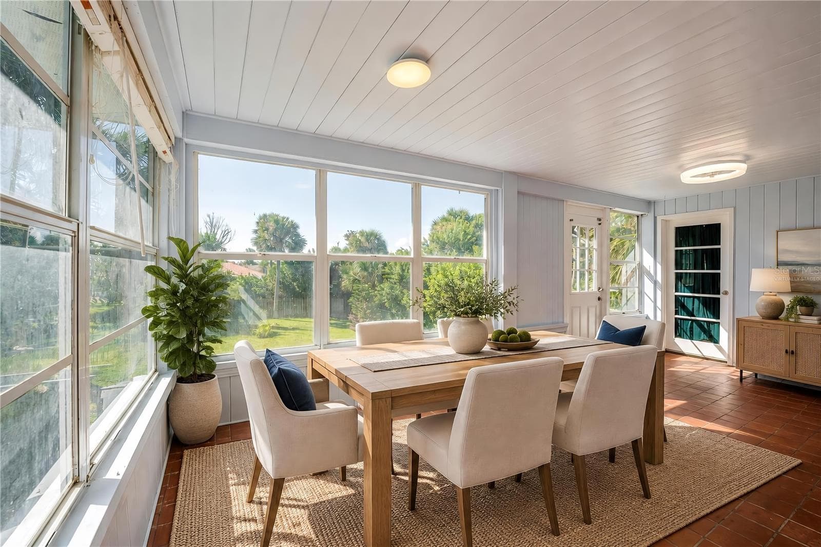 Virtually Staged - Breakfast nook featuring red tile floors, light-colored wood paneling, flush-mount ceiling lights, and wide windows overlooking the green backyard. Exterior glass doors provide easy access to the lawn and surrounding grounds. - 128 MINERVA RD, DAYTONA BEACH