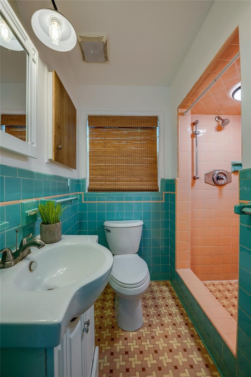 Vibrant vintage-style bathroom featuring turquoise wall tile, patterned geometric floor tile, and a matching pink-tiled walk-in shower stall. A white pedestal-style vanity with modern fixtures adds functional contrast. - 128 MINERVA RD, DAYTONA BEACH