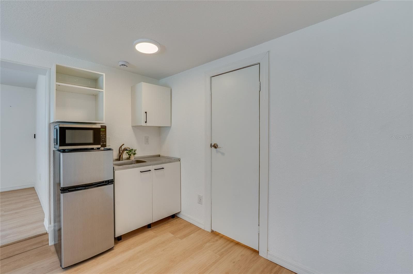 Functional kitchenette area equipped with a compact stainless-steel mini refrigerator, microwave, and white cabinetry with a sink. Clean white walls and warm LVP floors complete the modern, low-maintenance design. - 128 MINERVA RD, DAYTONA BEACH