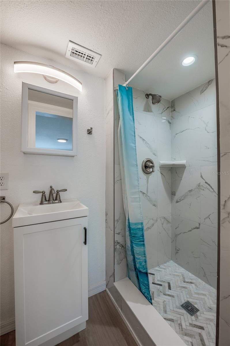 Modernized bathroom featuring a white single-sink vanity with modern hardware and a wall-mounted mirror. The walk-in shower is styled with marble-patterned wall tile and intricate herringbone floor tiling. - 128 MINERVA RD, DAYTONA BEACH