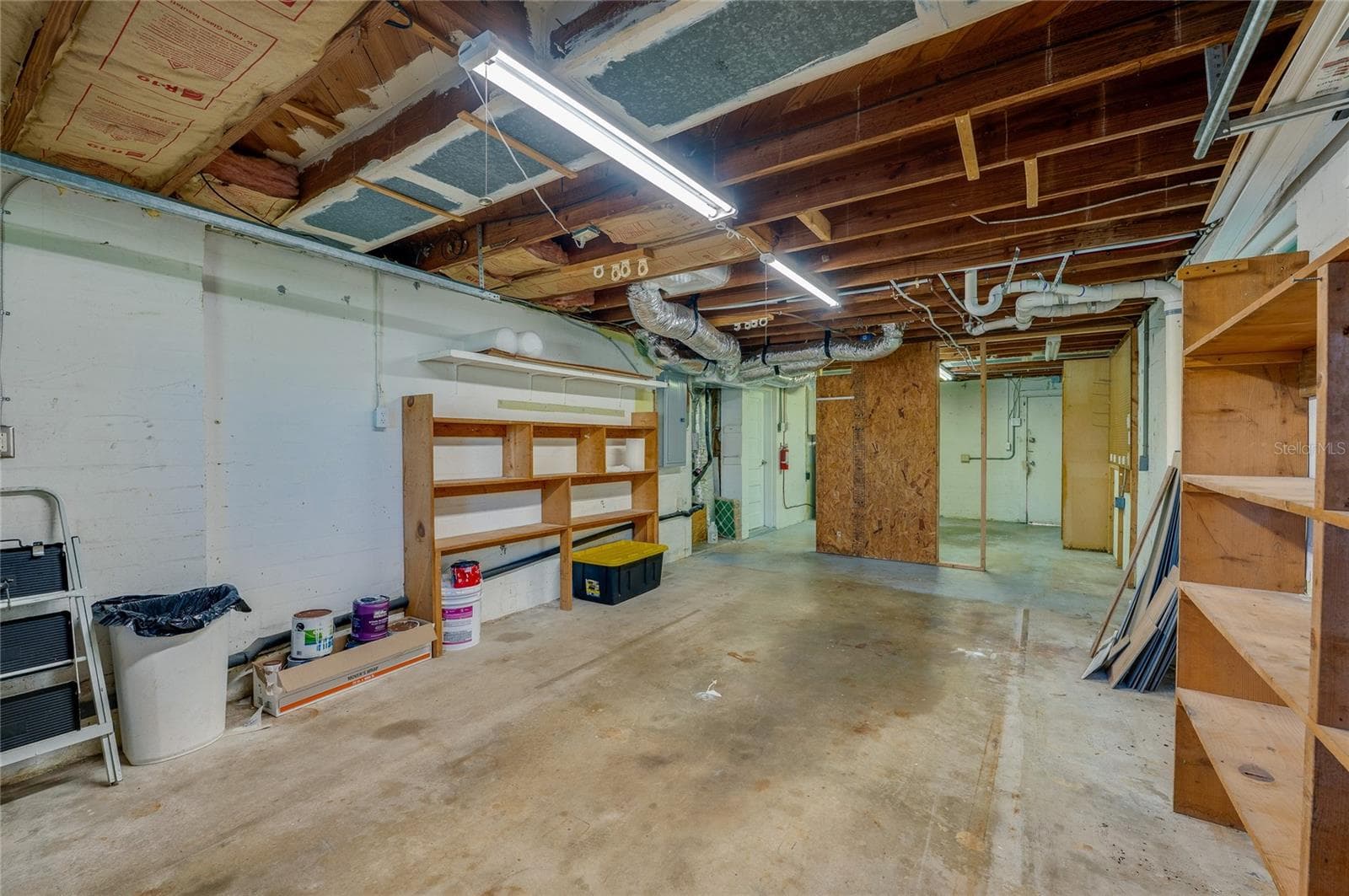 Expansive lower-level workshop or storage area featuring concrete flooring, exposed ceiling joists with overhead strip lighting, and built-in wooden shelving units. - 128 MINERVA RD, DAYTONA BEACH
