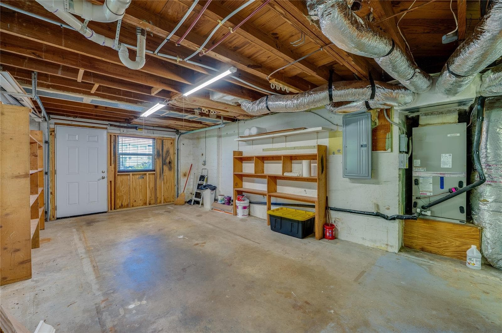 Functional utility room showcasing exposed overhead piping, ductwork, an HVAC unit, and an electrical panel. Built-in wooden shelving and a concrete slab floor offer practical storage space. - 128 MINERVA RD, DAYTONA BEACH