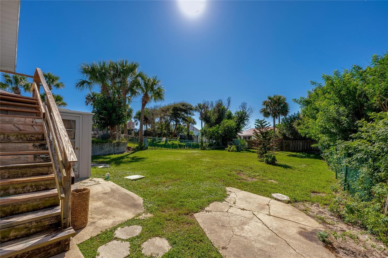 Spacious outdoor backyard featuring a grassy lawn, concrete patio stepping stones, and exterior wooden stairs leading up the residence. Lush tropical foliage and palm trees border the sunny yard. - 128 MINERVA RD, DAYTONA BEACH