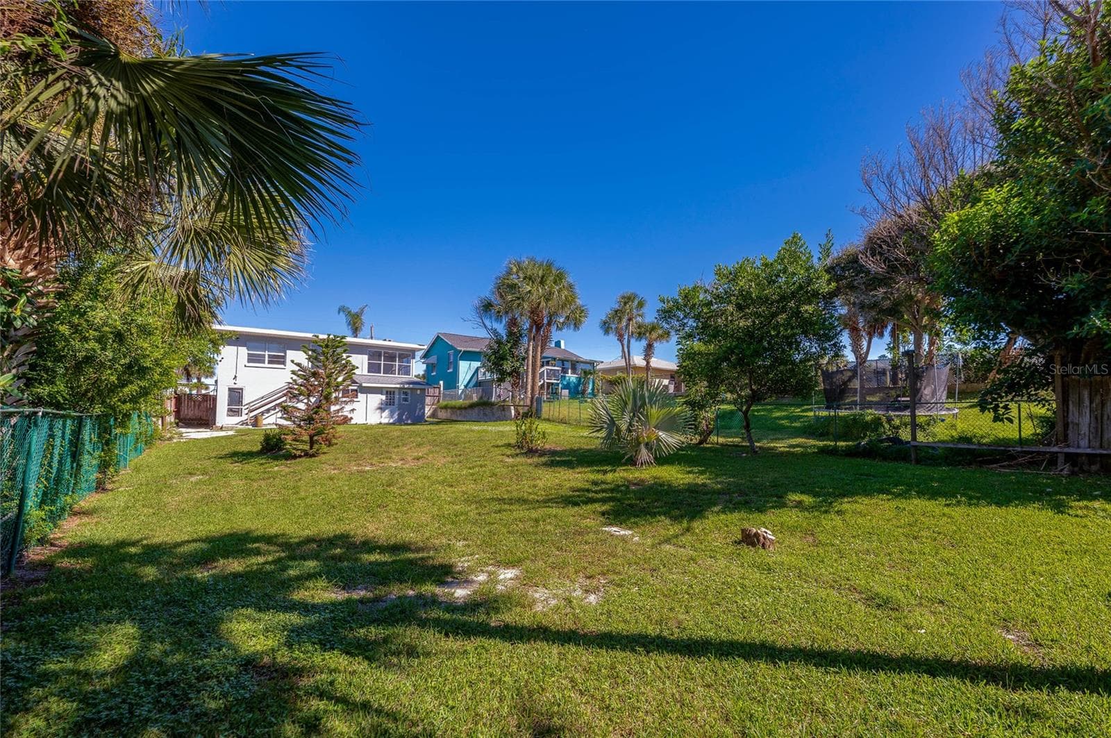 Expansive grassy backyard shaded by mature palm trees and green landscaping under a clear blue sky. The fenced perimeter offers privacy and ample room for outdoor recreation. - 128 MINERVA RD, DAYTONA BEACH