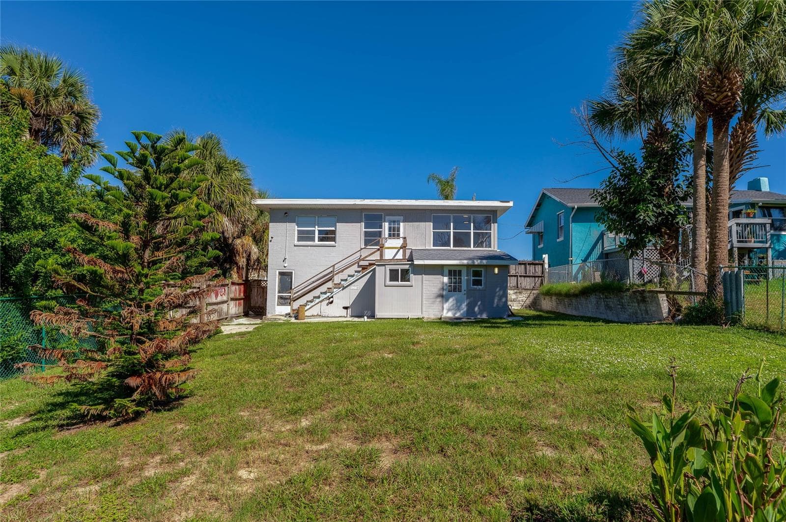 Rear view of a two-story home featuring an upper-level exterior staircase and a spacious grassy backyard. Vibrant palm trees and surrounding greenery enhance the coastal outdoor setting. - 128 MINERVA RD, DAYTONA BEACH