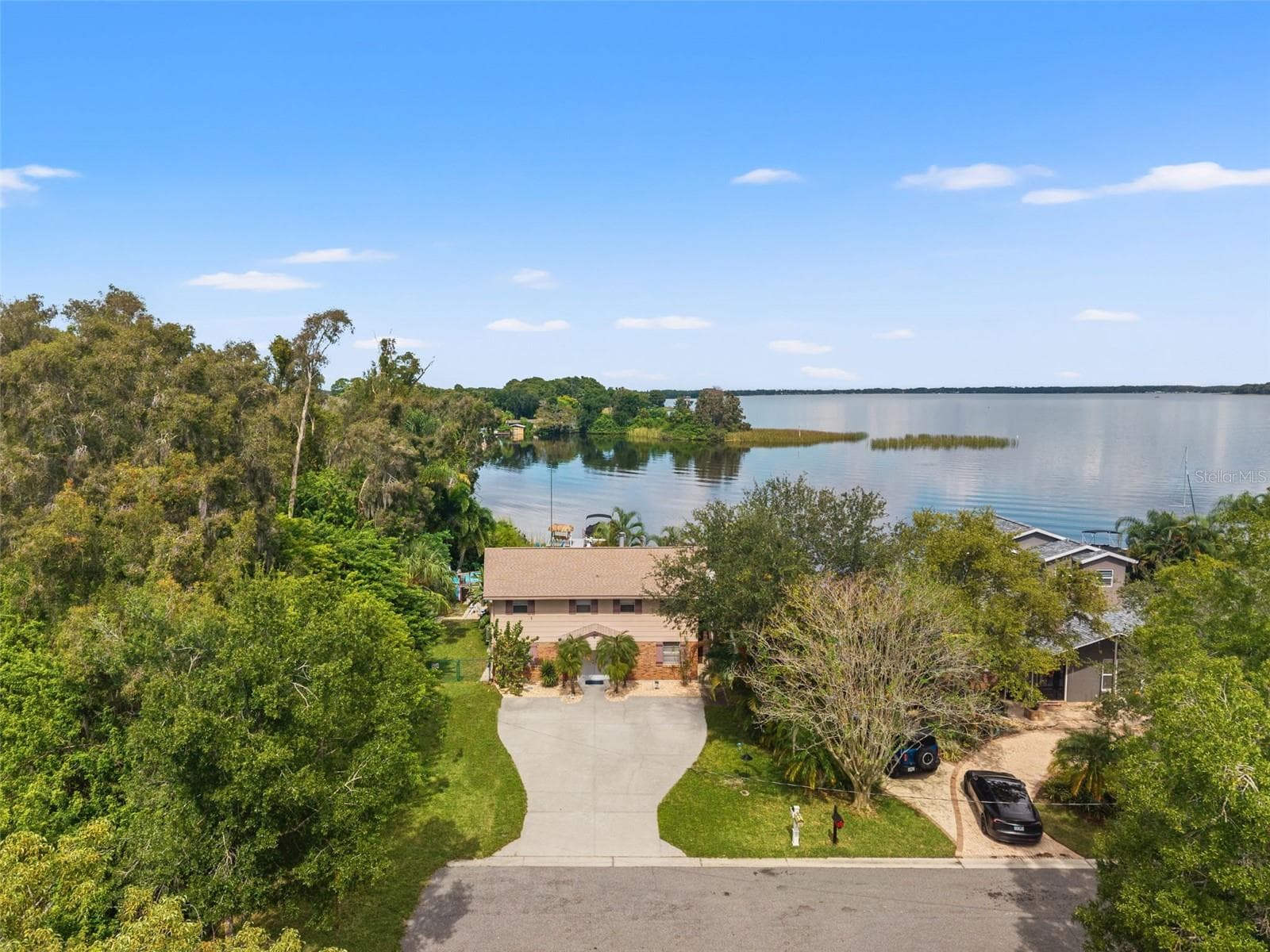 This aerial perspective captures the home's setting along a quiet residential street, surrounded by mature trees with the expansive lake stretching out behind it. The elevated home enjoys a generous driveway, lush greenery, and a waterfront location that makes boating and outdoor living part of everyday life. - 13 FRESHWATER, PALM HARBOR
