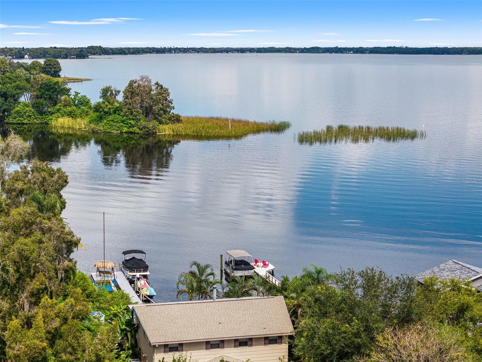An elevated perspective highlights the property's dock, pontoon boat lift, and thatched-roof seating area. Beyond the shoreline, expansive open water stretches toward the distant tree line, offering an inviting setting for boating, fishing, and enjoying the outdoors. - 13 FRESHWATER, PALM HARBOR