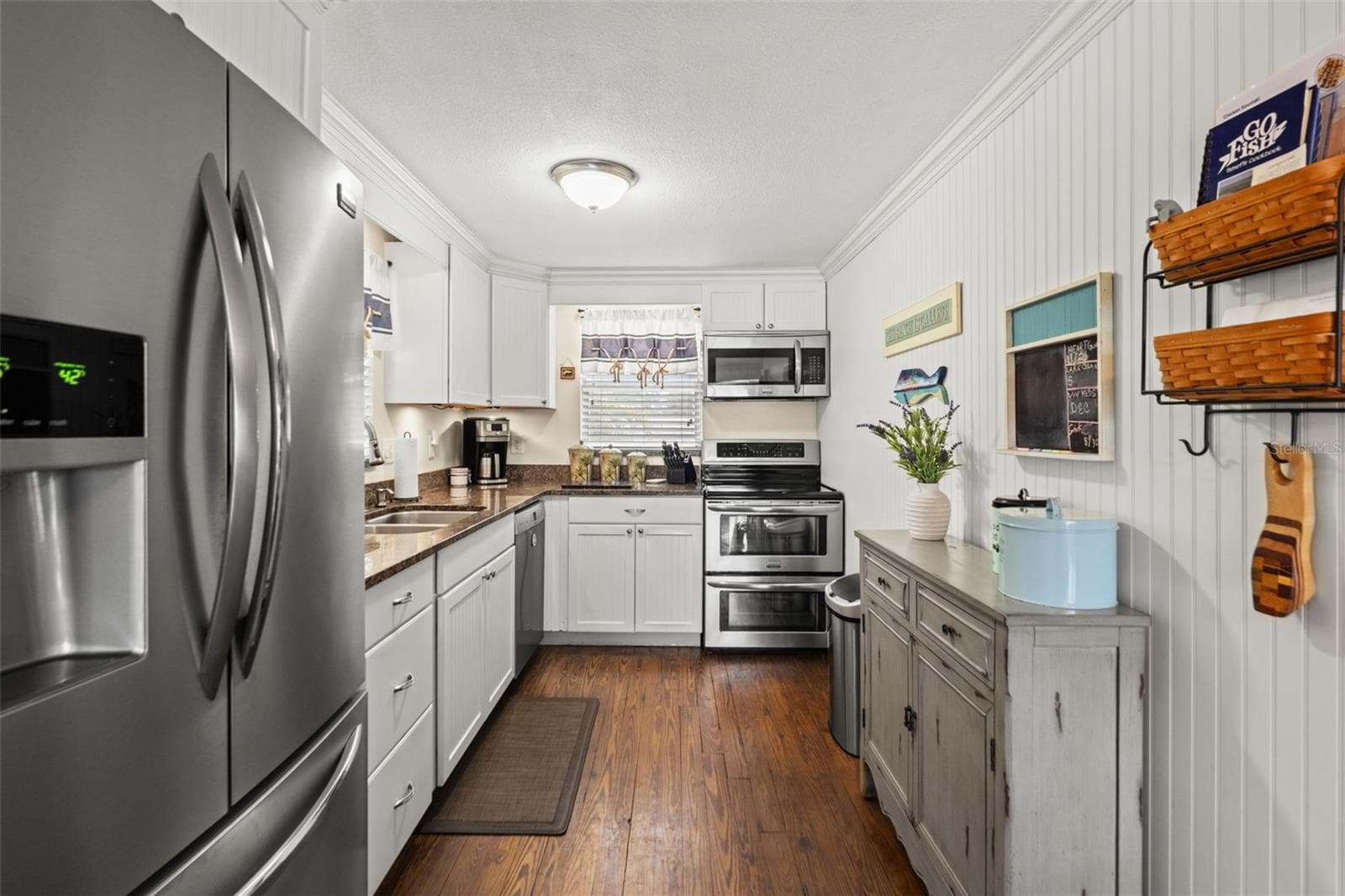 This inviting kitchen combines white cabinetry, stainless steel appliances, wood flooring, and warm-toned countertops. Crown molding and beadboard-style wall detailing add character, while the galley layout keeps everything conveniently within reach. - 13 FRESHWATER, PALM HARBOR