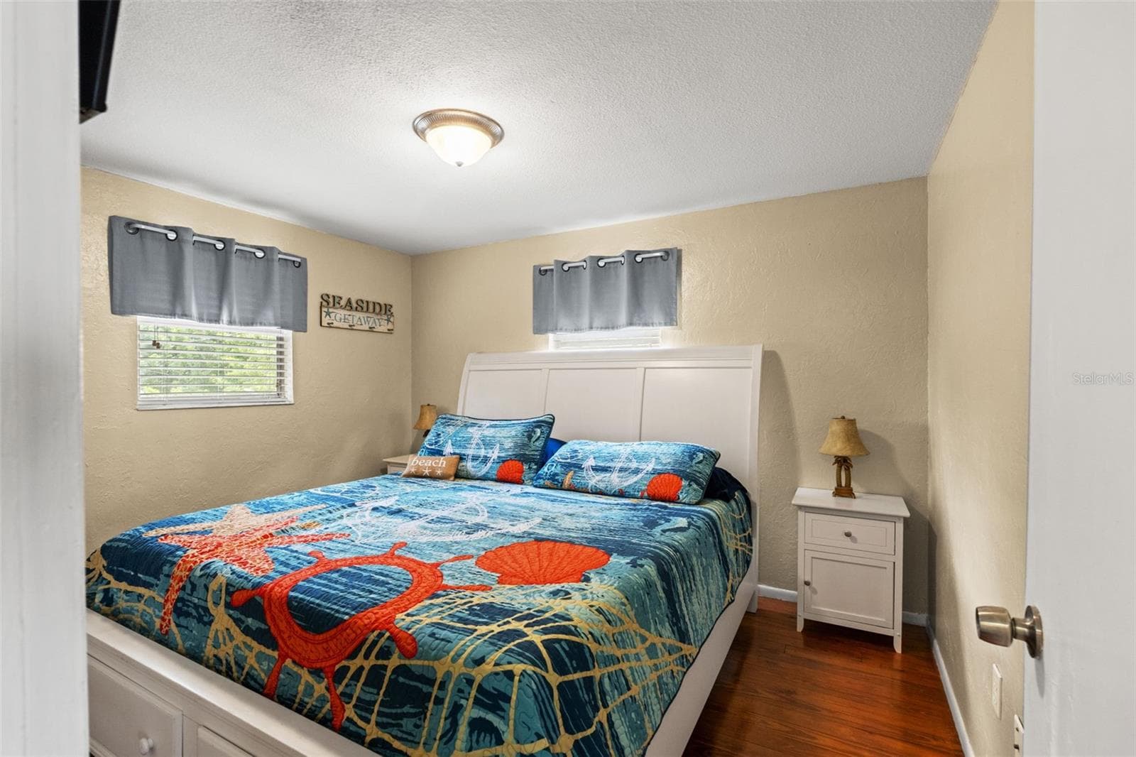 A comfortable bedroom with warm wood flooring, neutral walls, and two windows providing natural light. The room accommodates a generously sized bed with nightstands, while its coastal-inspired décor complements the home's relaxed waterfront setting. - 13 FRESHWATER, PALM HARBOR