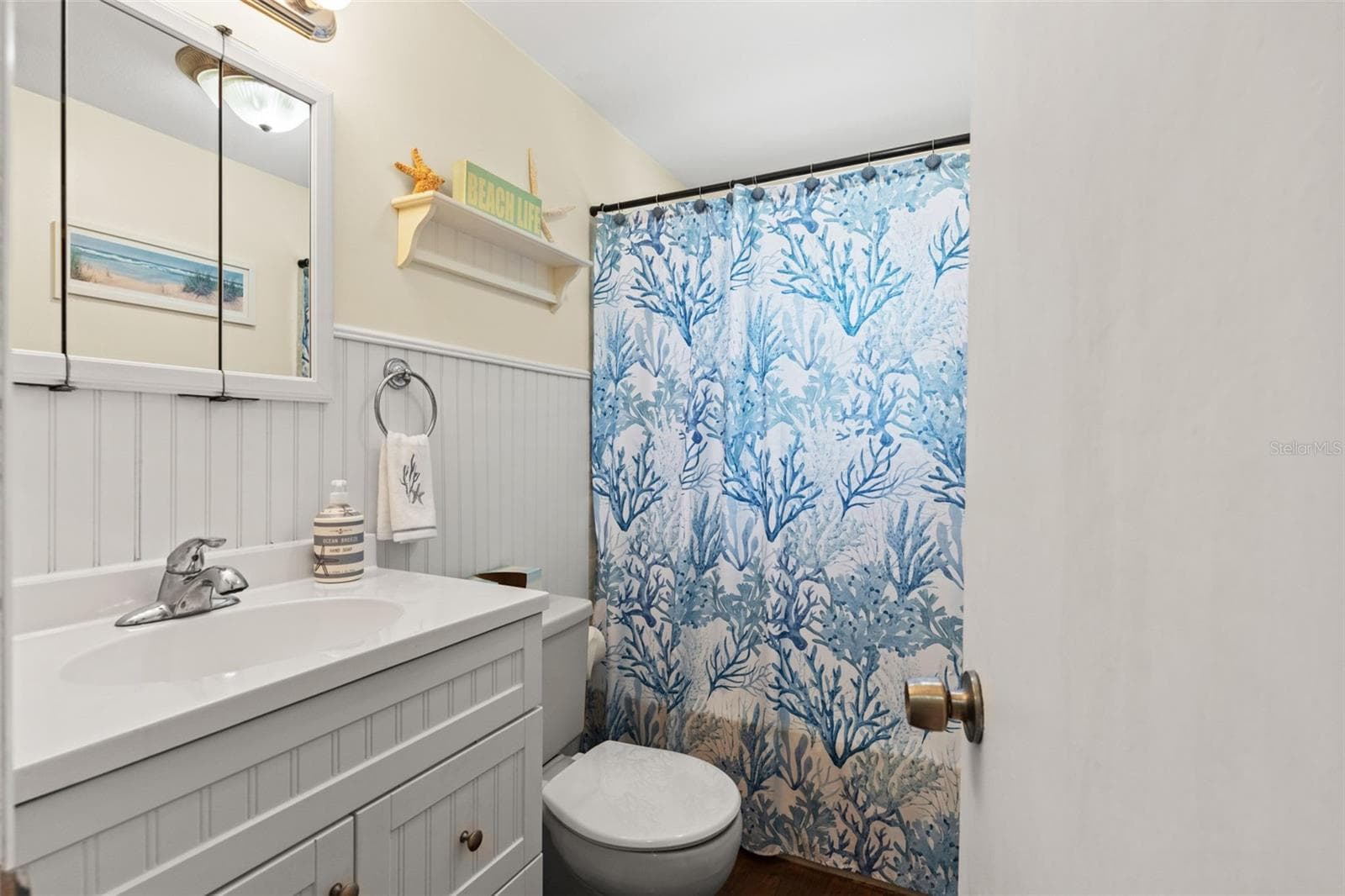 Coastal charm continues into this full bathroom, featuring a white vanity with decorative beadboard-style detailing, a mirrored medicine cabinet, and a tub/shower combination. White wainscoting and cheerful blue accents complete the beach-inspired look. - 13 FRESHWATER, PALM HARBOR