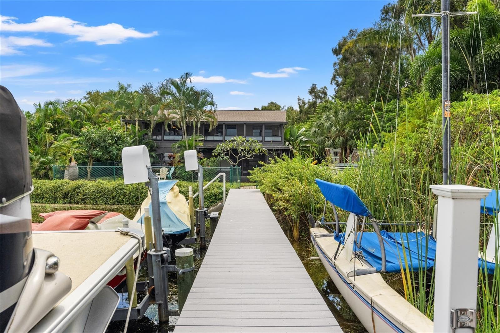 A view from the dock looking back toward the home showcases its elevated design, tropical landscaping, and waterfront setting. The long dock connects the backyard to the lake, with boat and watercraft lifts providing convenient access for outdoor recreation. - 13 FRESHWATER, PALM HARBOR