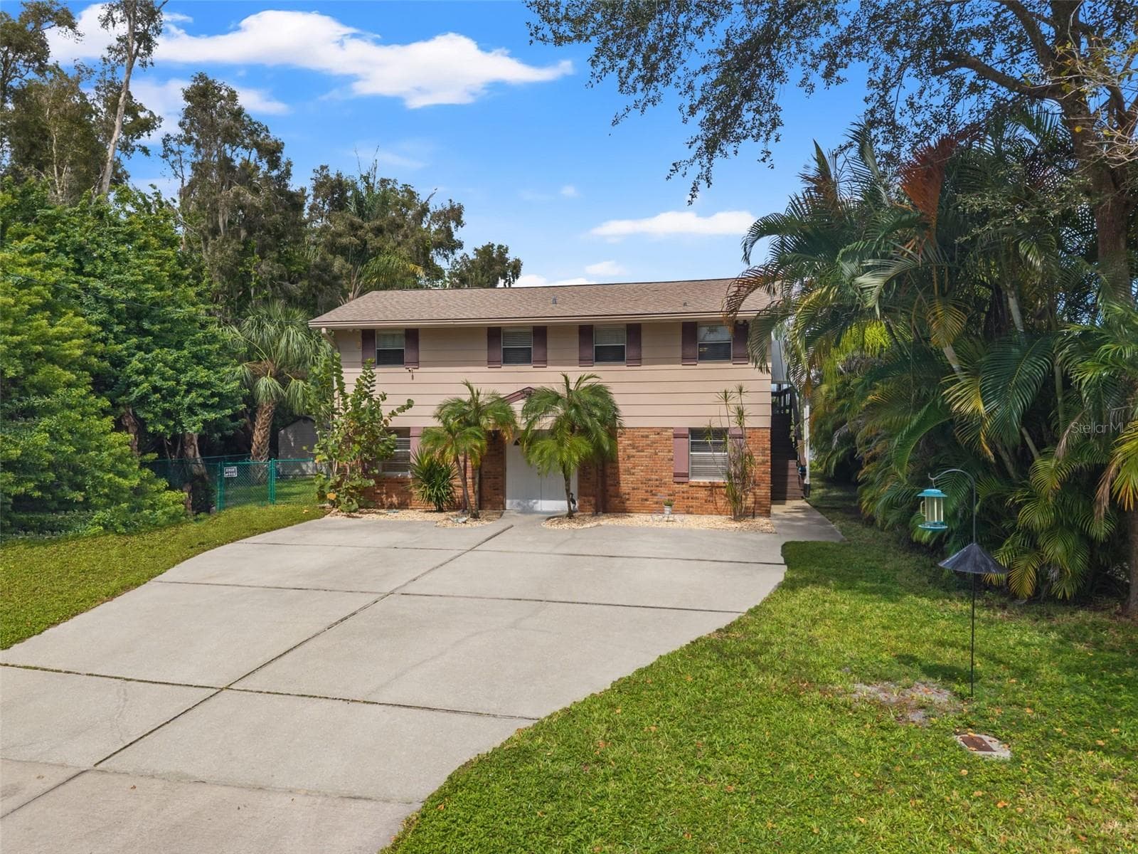Set among mature trees and tropical landscaping, this elevated home features a brick-accented lower level, a broad concrete driveway, and an upper-level living area. The spacious driveway offers generous parking, while the exterior staircase provides access to the elevated portion of the home. - 13 FRESHWATER, PALM HARBOR