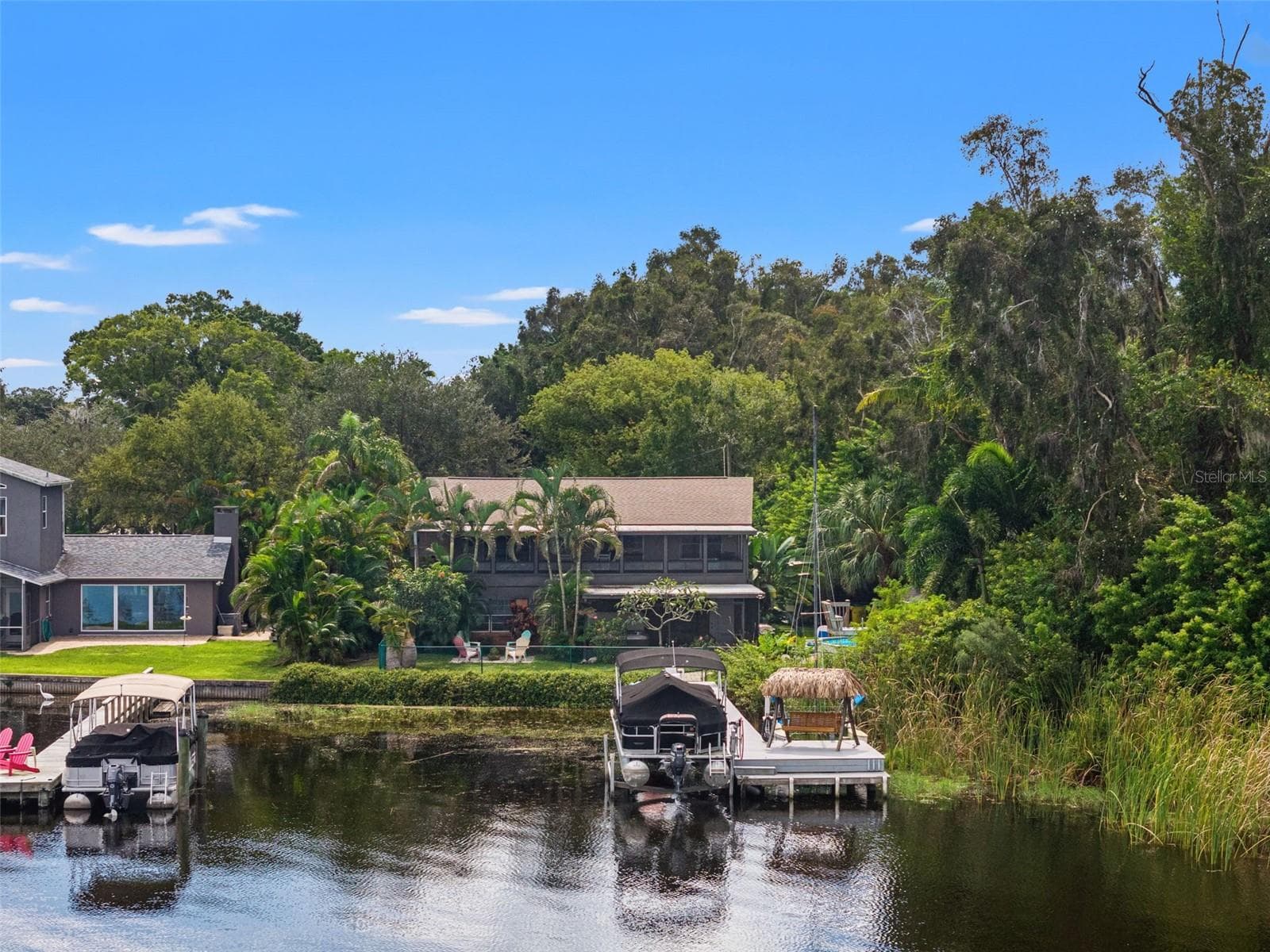 The home's waterfront setting is particularly appealing from this perspective. A private dock with a pontoon boat lift and thatched-roof swing sits along the shoreline, while dense vegetation borders one side of the property, creating a sense of separation from neighboring homes. - 13 FRESHWATER, PALM HARBOR