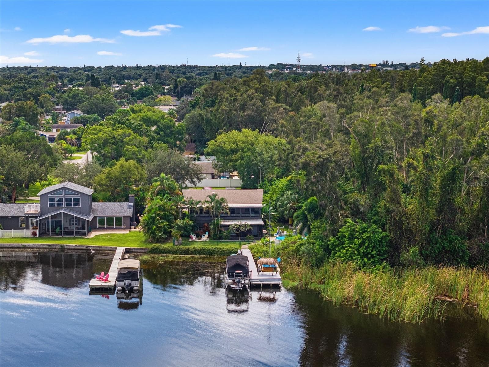 Looking toward the home from the water, this aerial photograph showcases the dock, pontoon boat lift, and shaded swing area. Tropical palms and mature trees surround the elevated residence, while natural shoreline vegetation contributes to the property's peaceful waterfront atmosphere. - 13 FRESHWATER, PALM HARBOR