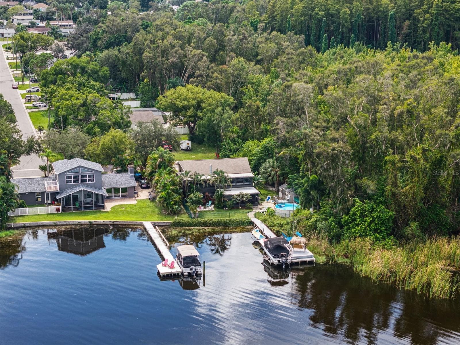This aerial photograph brings together several of the property's standout features: an elevated home, above-ground backyard pool, private dock with pontoon boat lift, and shaded thatched-roof swing. The adjoining wooded area creates a natural backdrop, making the waterfront setting especially appealing. - 13 FRESHWATER, PALM HARBOR