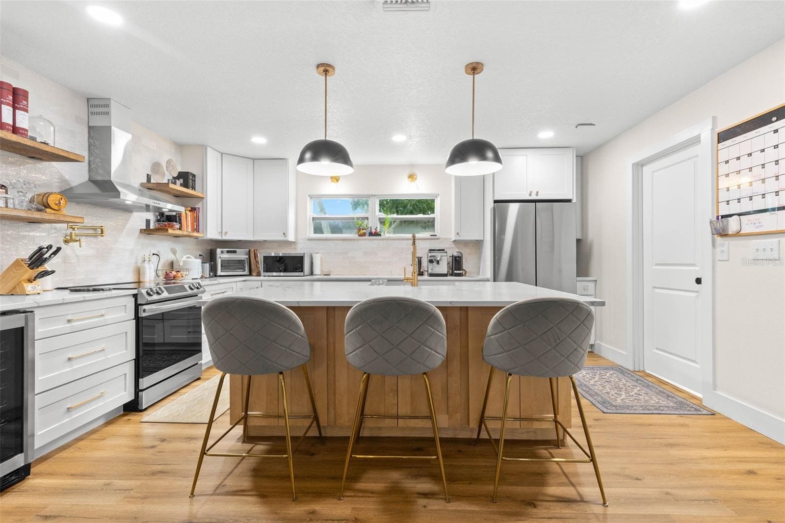 Beautifully updated and designed for both cooking and entertaining, this kitchen features an oversized quartz-topped center island with seating, white cabinetry with gold hardware, a warm wood-paneled island base, stainless steel appliances, and striking black dome pendant lights. Open shelving and a stylish tile backsplash complete the look. - 3013 GRAHAM LN, TAMPA